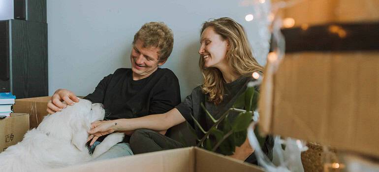 A man and a woman playing with their dog while sitting behind some moving boxes