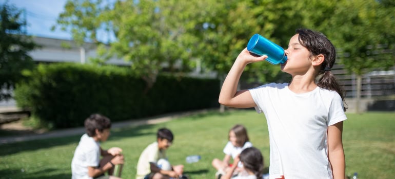 A girl in a park drinking water from her blue bottle.