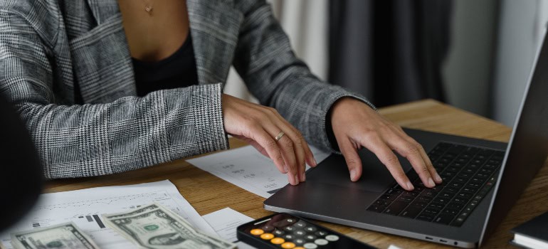 A woman doing budgeting by using her laptop, some papers, and a calculator on her phone.