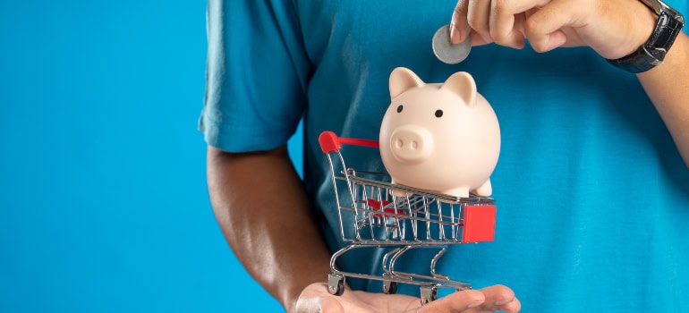 a person inserting a coin into a piggy bank placed above a tiny shopping trolley