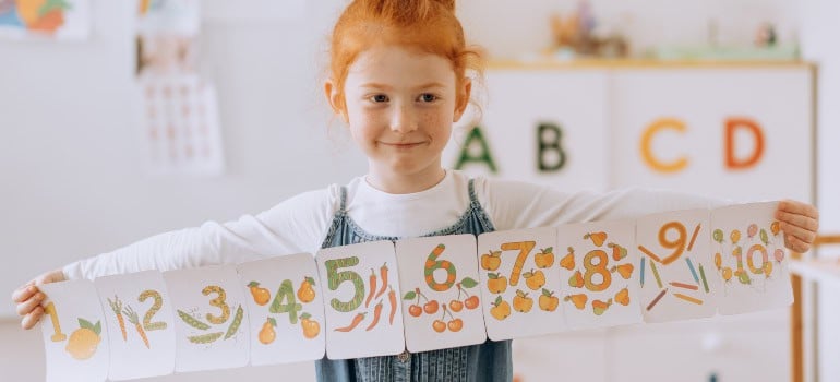 a little girl with ginger hair holding a banner with numbers and illustrations