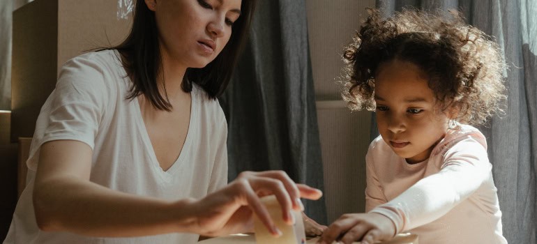 a woman and a child sealing a cardboard box with duct tape