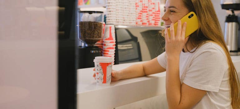 girl in a white shirt holds a cup of coffee and talks on the phone while smiling